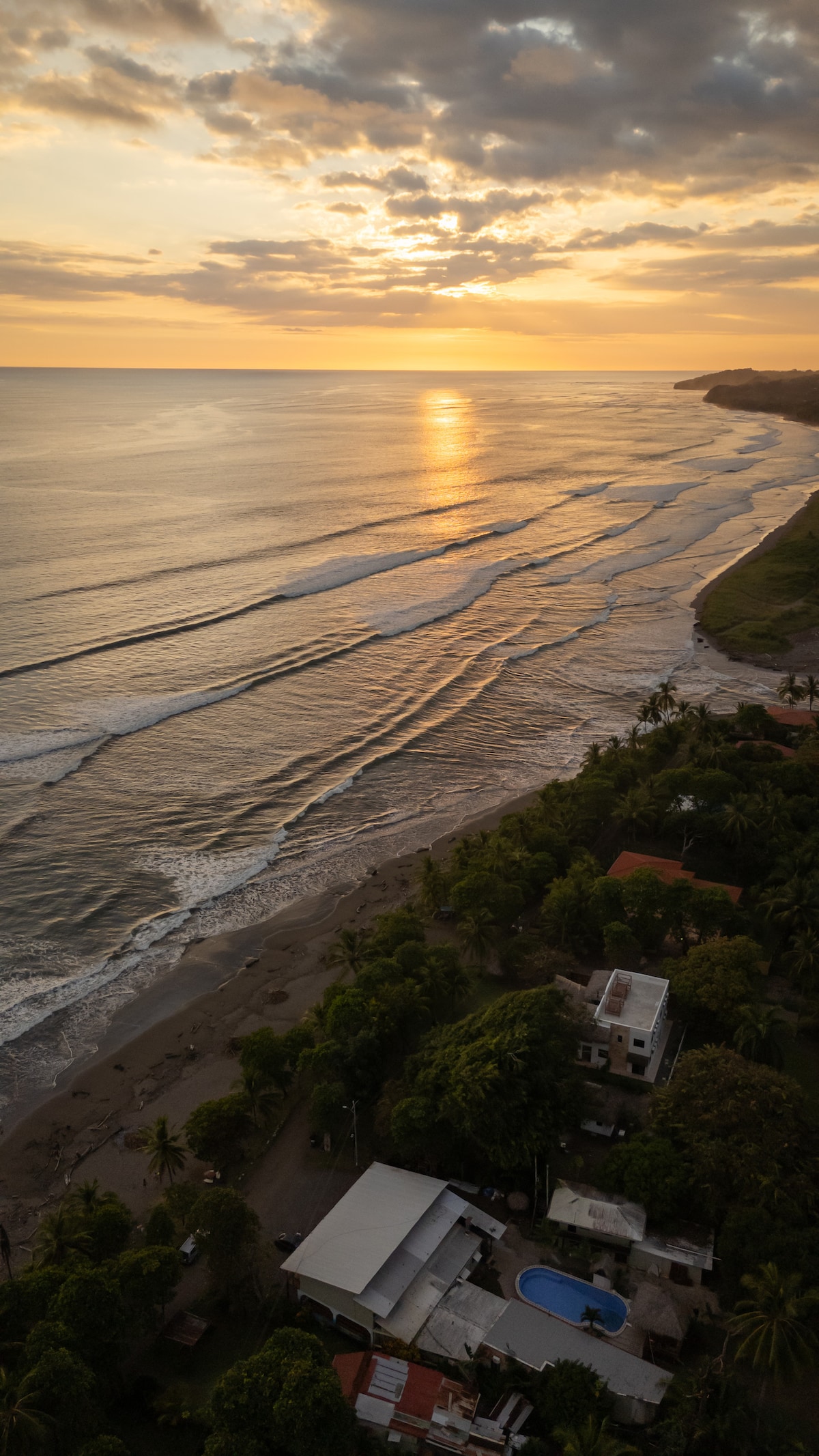 An aerial view captures a stunning sunset over the ocean, with golden sunlight reflecting on gentle waves. The tranquil beach is framed by lush greenery and gently sloping hills, while the property below features a swimming pool amidst palm trees and terracotta-roofed homes.