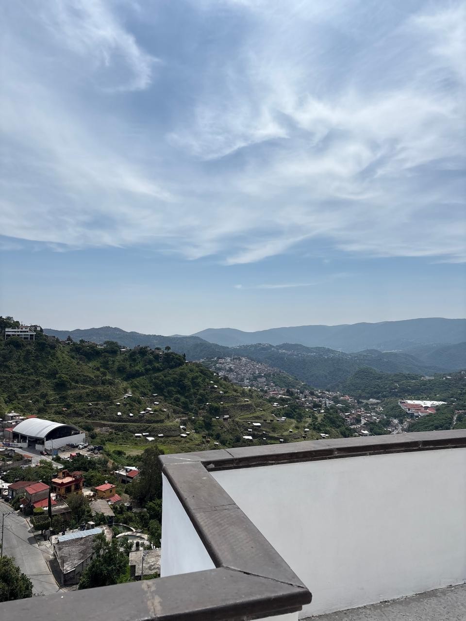 A panoramic view captures rolling green hills under a bright blue sky, with wisps of clouds scattered above. In the distance, a town is nestled among the mountains, showcasing a harmonious blend of nature and residential life.