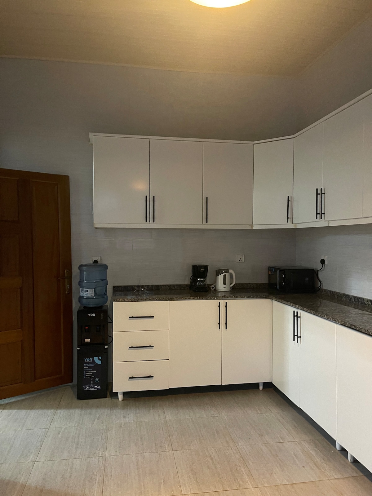 A modern kitchen area features white cabinetry and a granite countertop. Standard appliances such as a coffee maker and a microwave are present. A water dispenser is situated next to the cabinets, and natural light brightens the space through the room's layout.