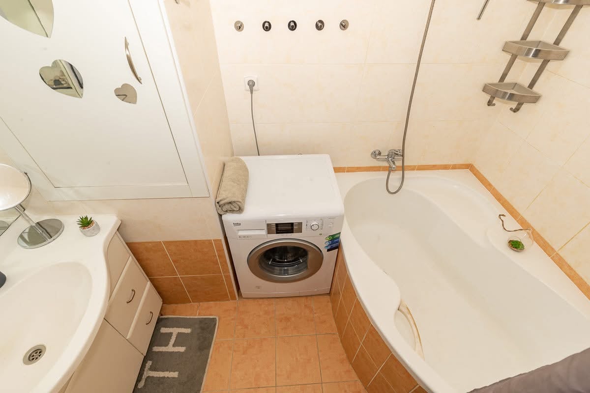 The bathroom features a compact layout with a comfortable bathtub and a separate washing machine. A mirrored cabinet is positioned above the sink, with a small decorative plant on the countertop. Neutral tones and minimalist design elements create a functional space.