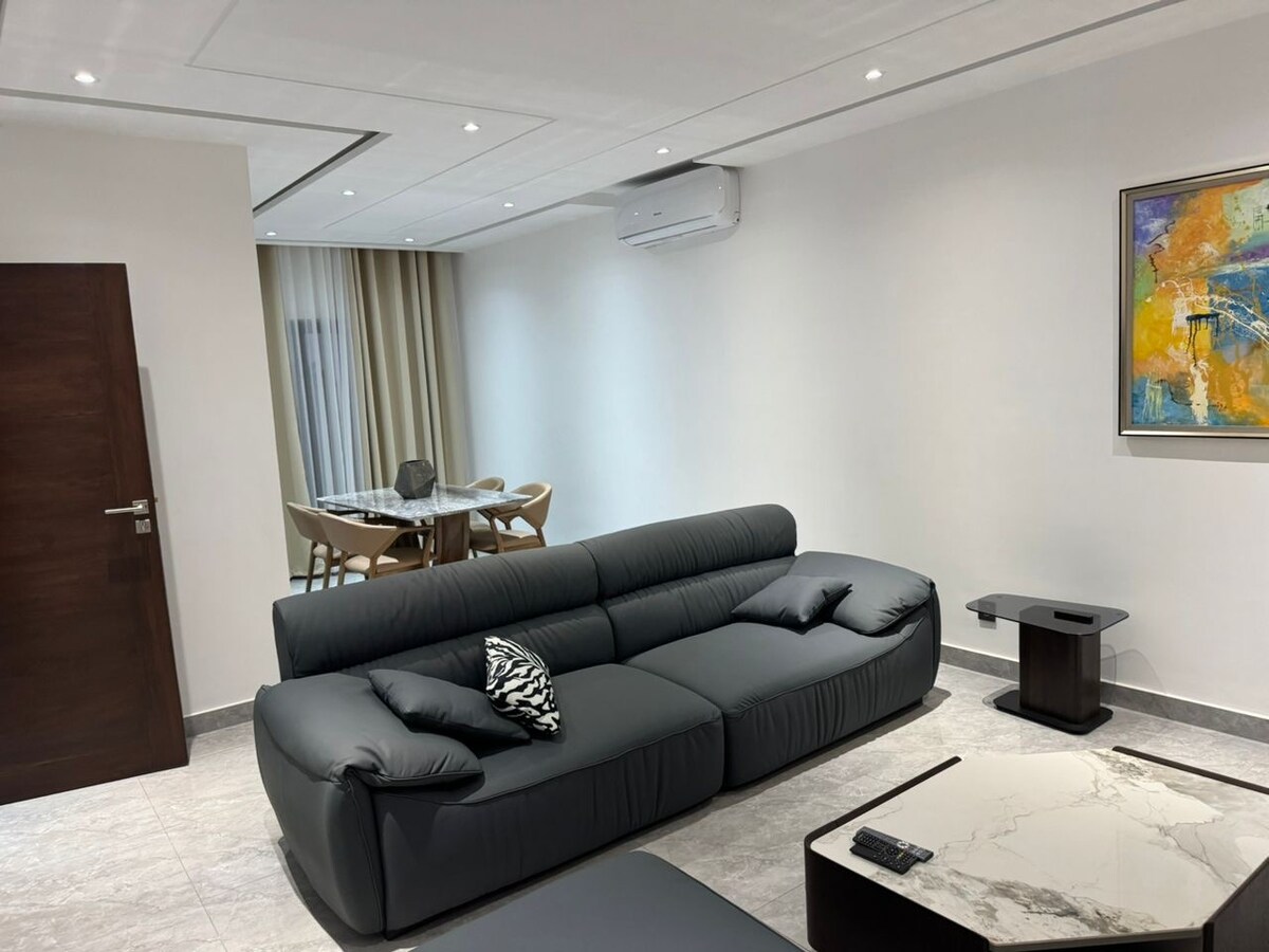 A contemporary living area features a dark sectional sofa with cushions, paired with a sleek coffee table. A dining table with light wood chairs is visible in the background, alongside large windows dressed with soft drapes. An artwork adds a pop of color to the wall.