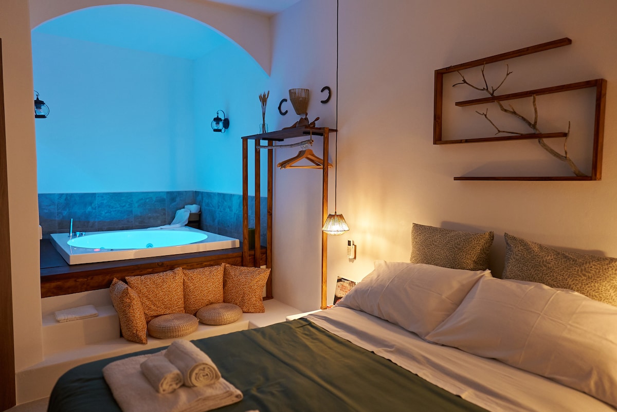 The room features a spacious bed with neutral-toned linens. A relaxing area is highlighted by a hot tub, softly illuminated in blue, located in the background. Cozy cushions provide seating alongside a wooden rack and decorative wall elements.