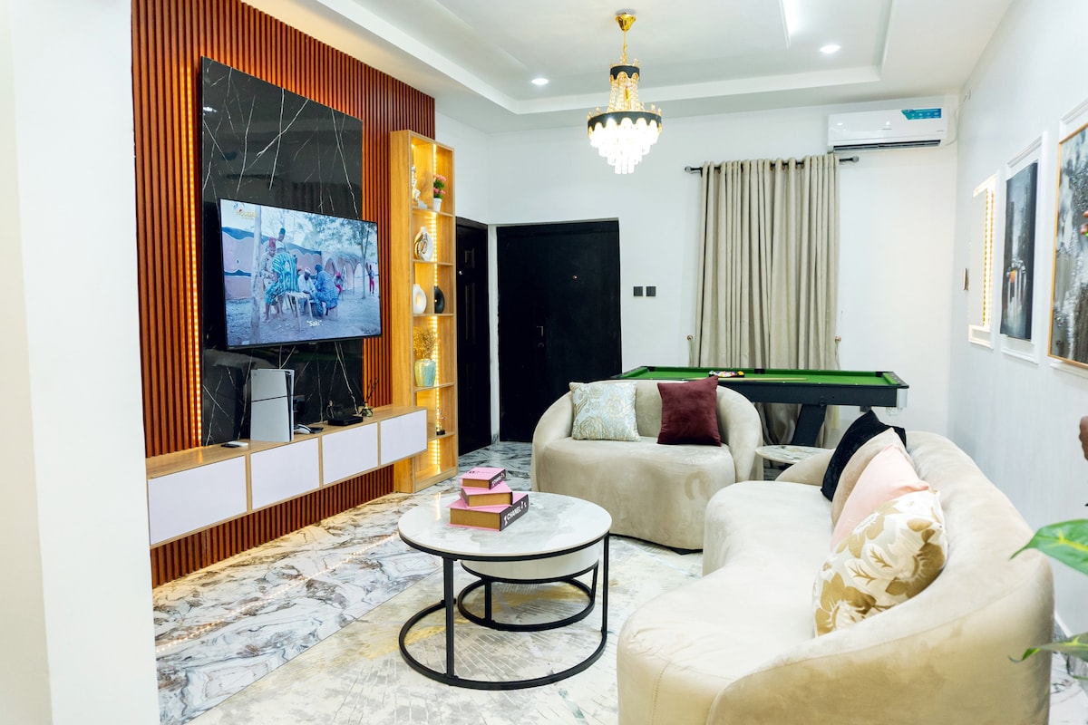 A modern living room showcases a large television mounted on a stylish feature wall, complemented by custom shelving. A cozy sectional sofa and circular coffee table create a comfortable seating area, while a pool table is positioned in the background.