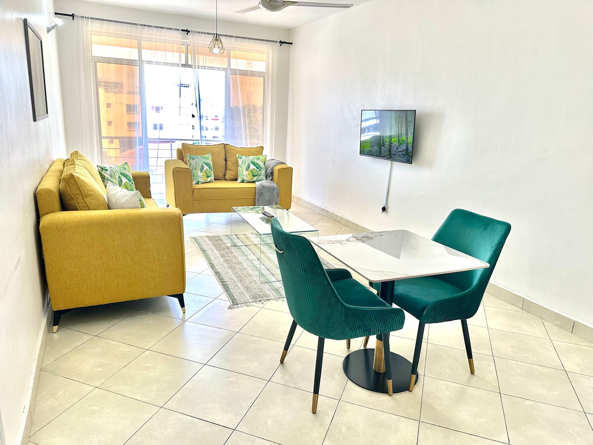 A bright living area showcases a yellow sofa adorned with patterned cushions, complemented by a glass coffee table. A dining nook with teal chairs and a marble table resides alongside a wall-mounted television, with natural light filtering through large windows.