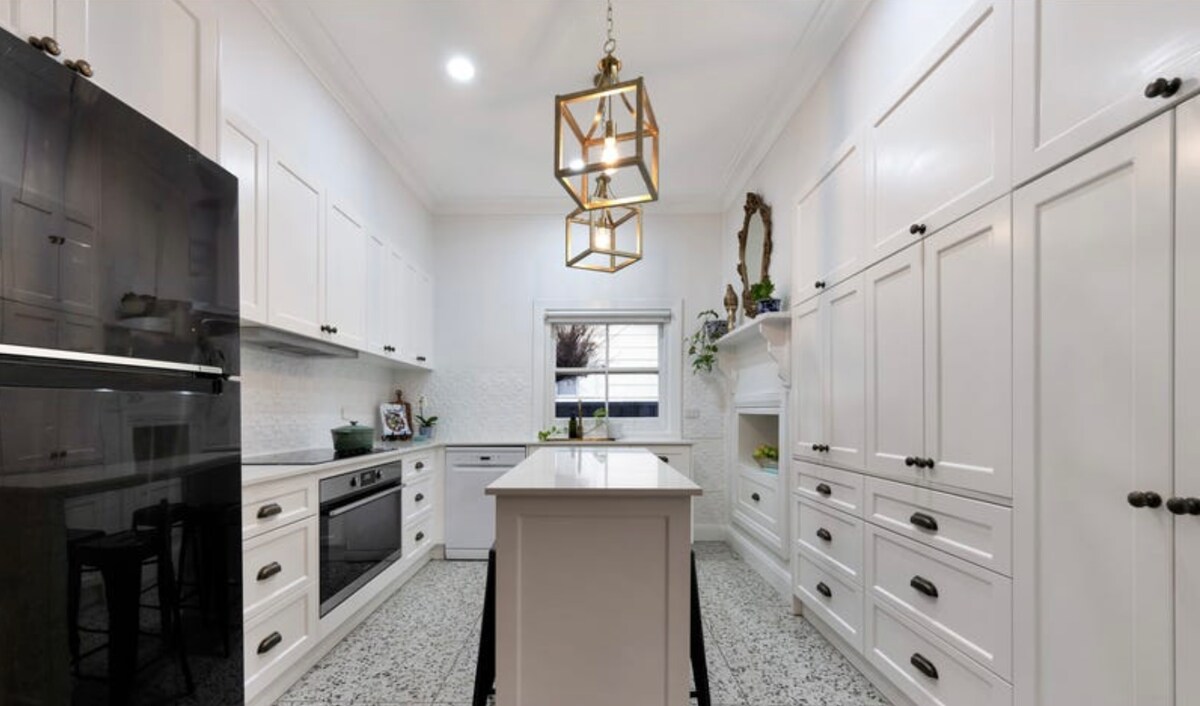 A modern kitchen is characterized by white cabinetry and a central island. A large black refrigerator stands nearby, while sleek appliances are integrated into the cabinetry. Natural light filters through a window, and a pendant light hangs above the island, adding a subtle focal point to the space.