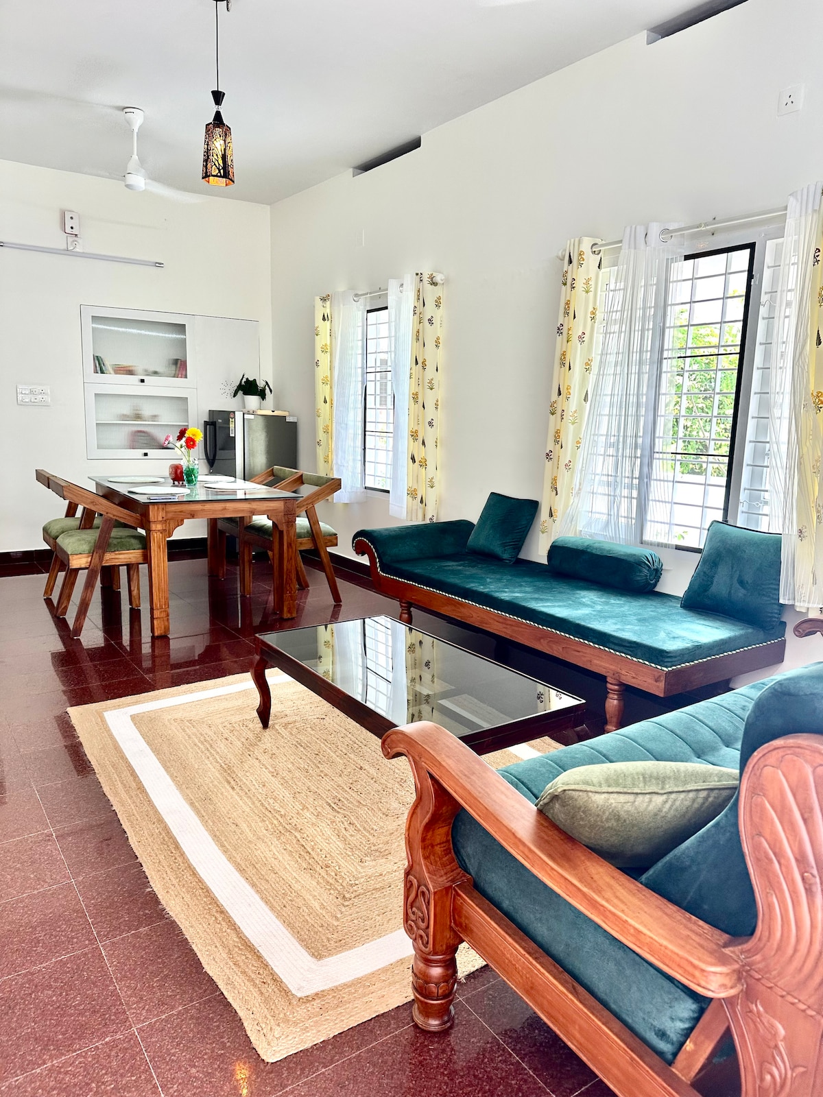 The living area features a three-seater sofa and a diwan cot with cushions, accompanied by a wooden dining table with seating for four. Large windows allow natural light to fill the space, and a light-coloured area rug adds warmth to the polished floor.