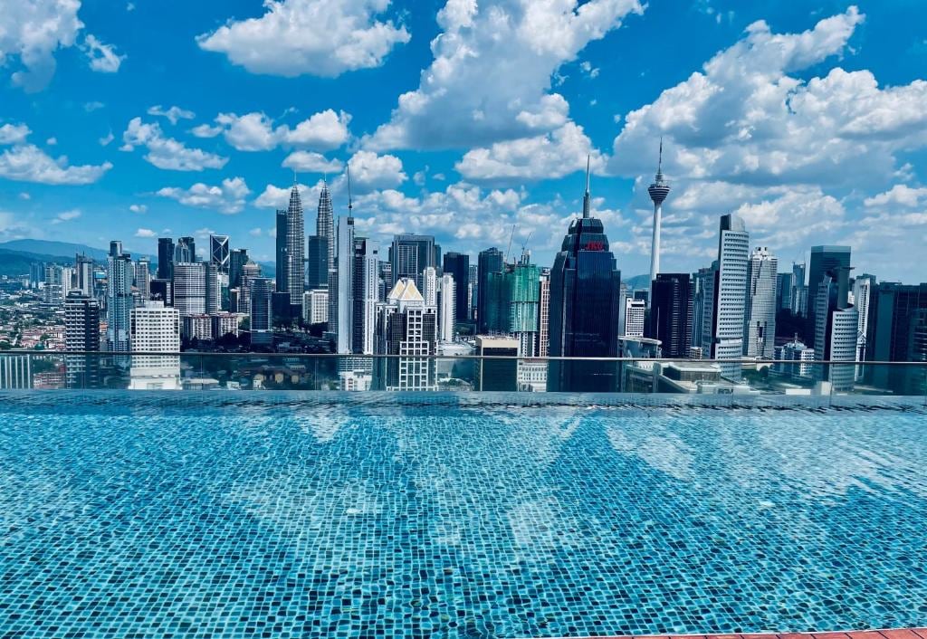 A shimmering sky pool is visible, its surface reflecting the bright blue sky dotted with clouds. The city skyline features a blend of modern skyscrapers, including the iconic Petronas Towers, creating a vibrant urban backdrop.