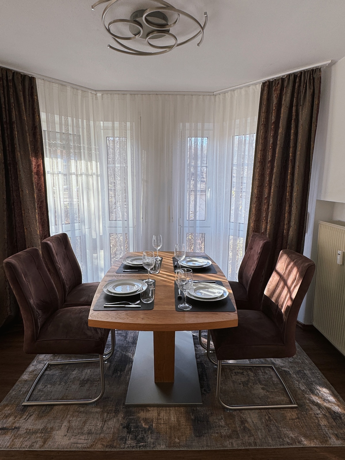 A modern dining area is illuminated by natural light from large windows, revealing a wooden table set for four. The space features upholstered chairs and elegant table settings, complemented by sheer curtains and a softly textured rug, creating an inviting dining atmosphere.