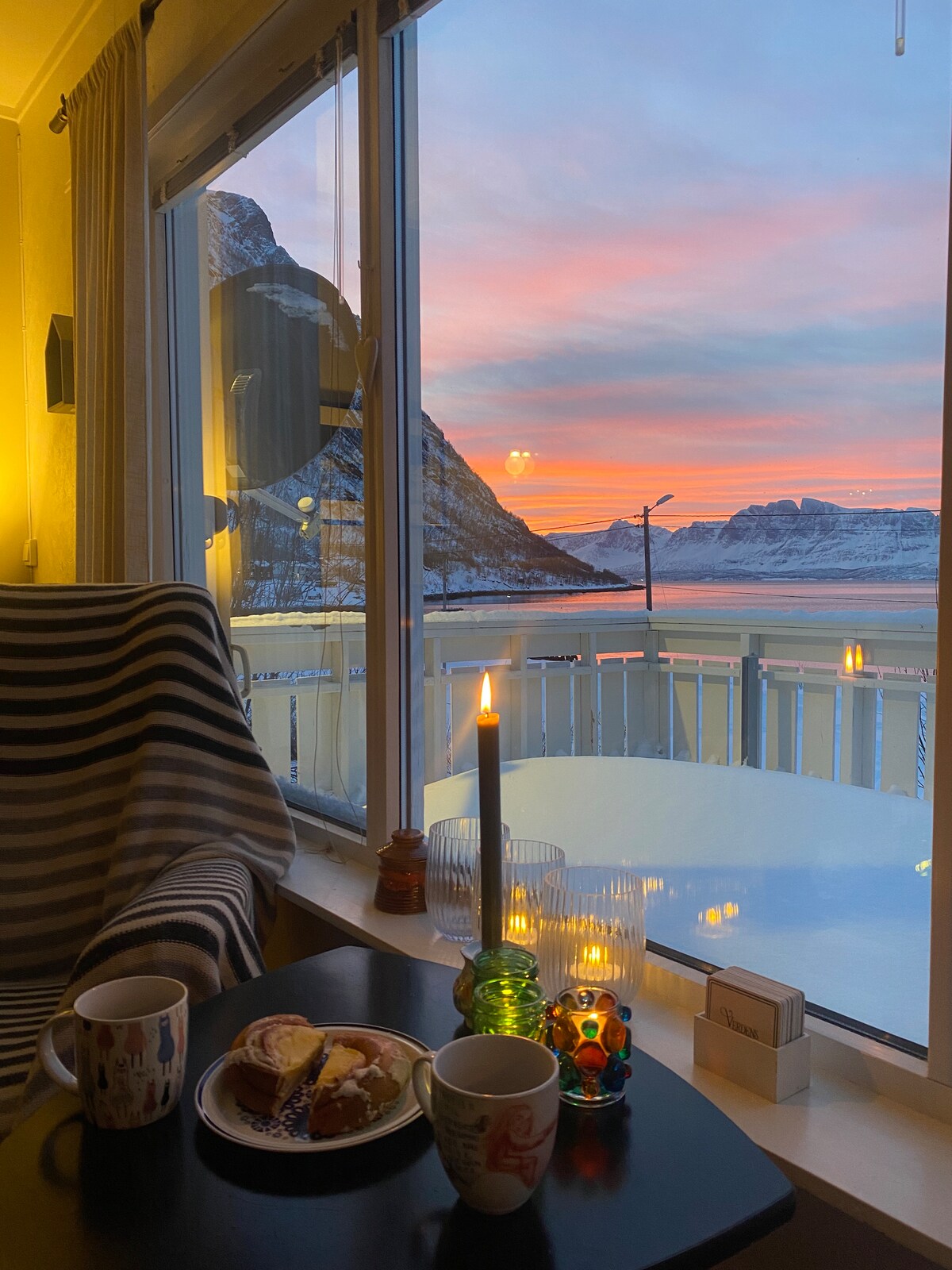 A cozy seating area is seen through large windows, showcasing a breathtaking sunset over the mountains. A table holds two mugs and a plate with pastries, accompanied by a lit candle and decorative glass holders, enhancing the tranquil ambiance.