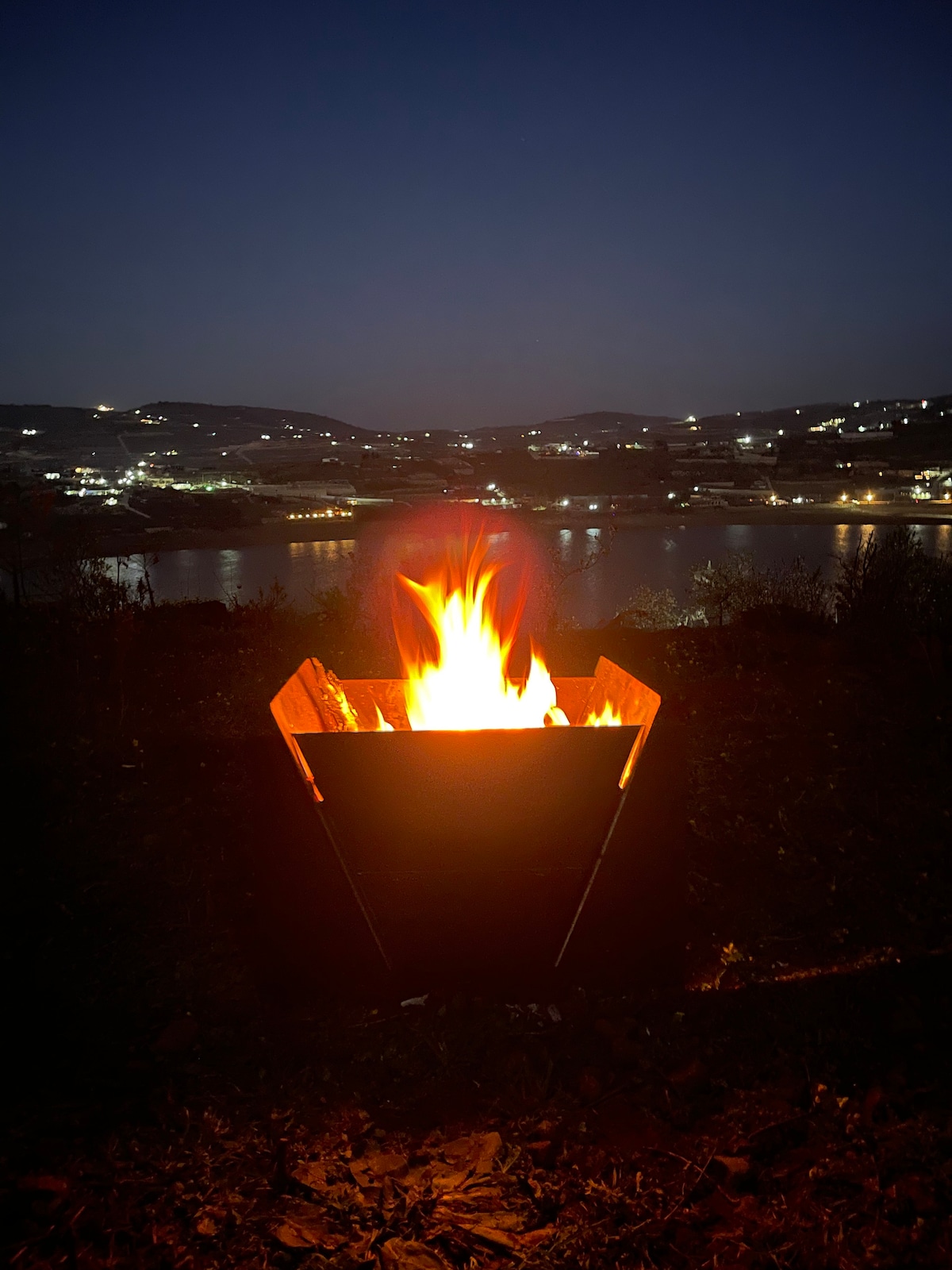 A fire pit is prominently featured, glowing warmly against the night. Beyond, a view of the calm water reflects distant lights from the surrounding landscape, creating a serene atmosphere under the night sky.