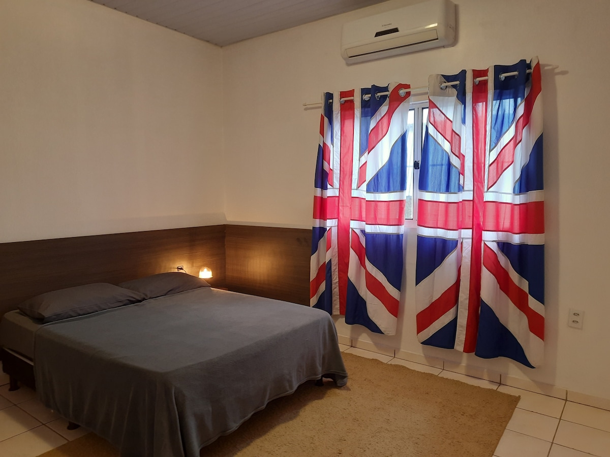 A spacious bedroom is furnished with a large bed covered in a simple gray blanket. The walls are complemented by curtains featuring a Union Jack design, framing a window that allows natural light to enter the room. An air conditioning unit is mounted above the bed.