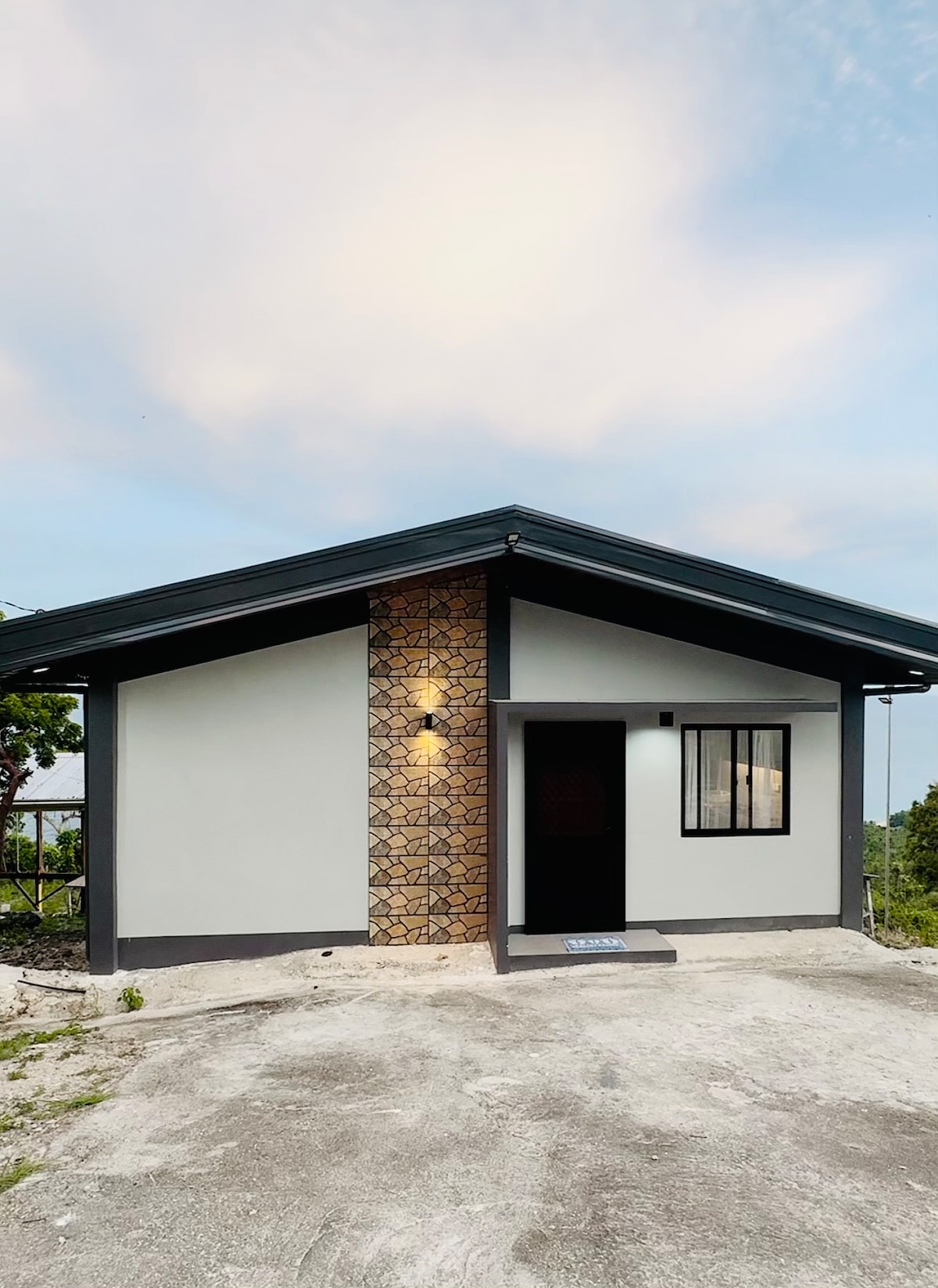 A modern single-story structure features a sloped roof and a decorative stone panel on one exterior wall. The entrance is marked by a dark door with a window beside it. Surrounding landscaping includes a gravel path and green foliage.