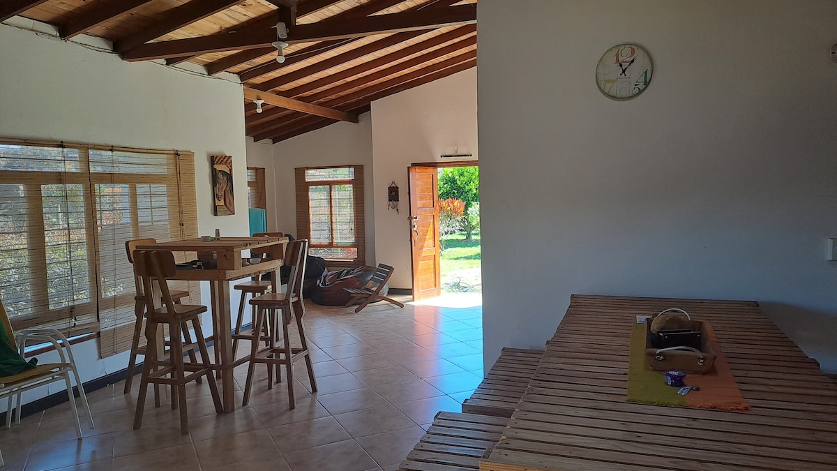 An open-plan living area features wooden beams and large windows, allowing natural light to fill the space. A high table with chairs is positioned centrally, while a comfortable lounge chair can be seen nearby. Doors lead to a garden with greenery outside.