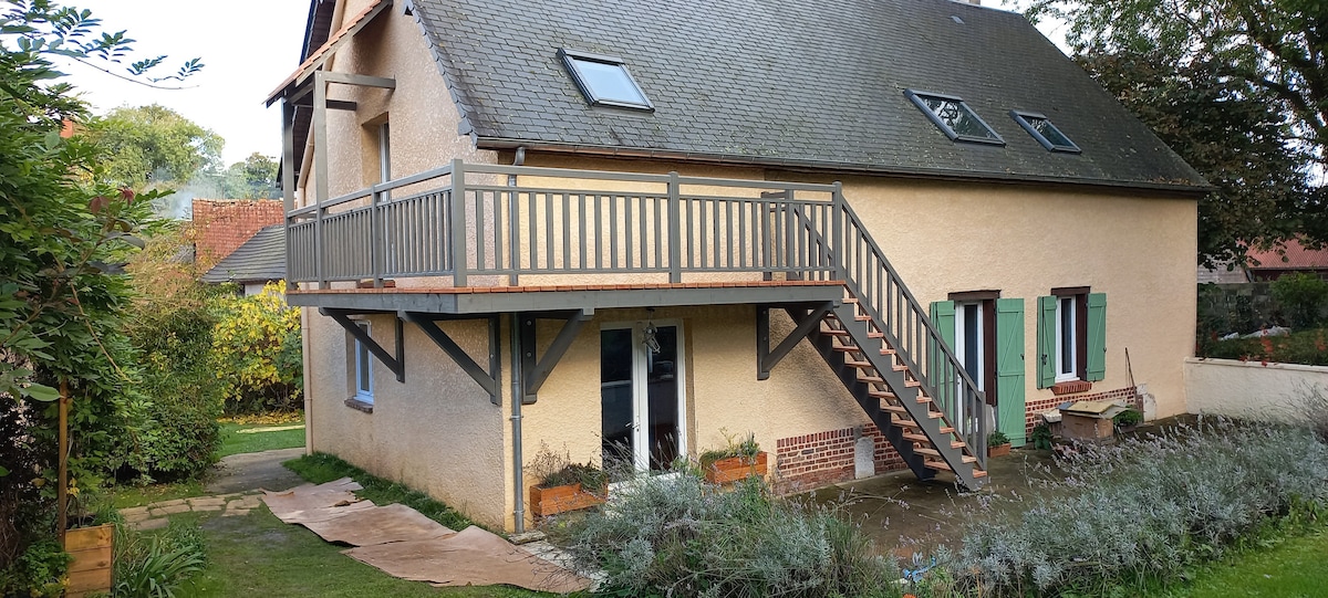 A two-story building features a balcony with a wooden railing and a set of stairs leading to the ground level. Green shutters frame several windows, enhancing the exterior's design. Lush greenery and garden beds adorn the area around the building.