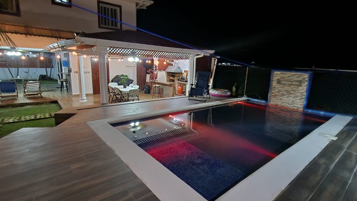 A modern outdoor area is highlighted by a private pool, which is illuminated in shades of red and blue. Adjacent, a covered seating space is furnished with comfortable chairs, and a grill area is visible in the background, surrounded by landscaped green spaces.