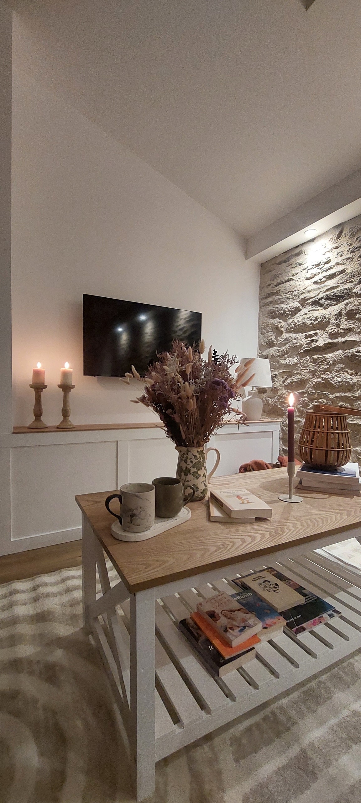 A cozy living space is highlighted by a wooden coffee table adorned with decorative items, including a vase of dried flowers and two mugs. A flat-screen TV is mounted on a white wall, while a textured stone accent wall adds depth. Soft lighting is provided by candles.