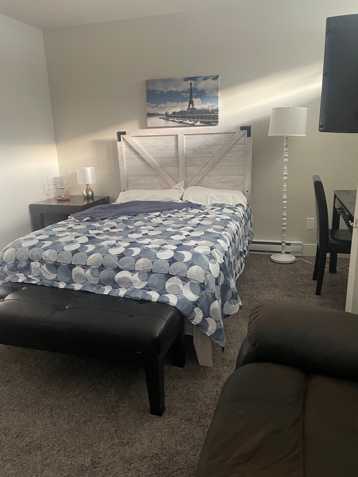 A cozy sleeping area features a bed with a light blue and white patterned blanket. Two bedside tables are positioned on either side, with a lamp on one. A seating area includes a dark leather chair, while a desk and television complete the space.