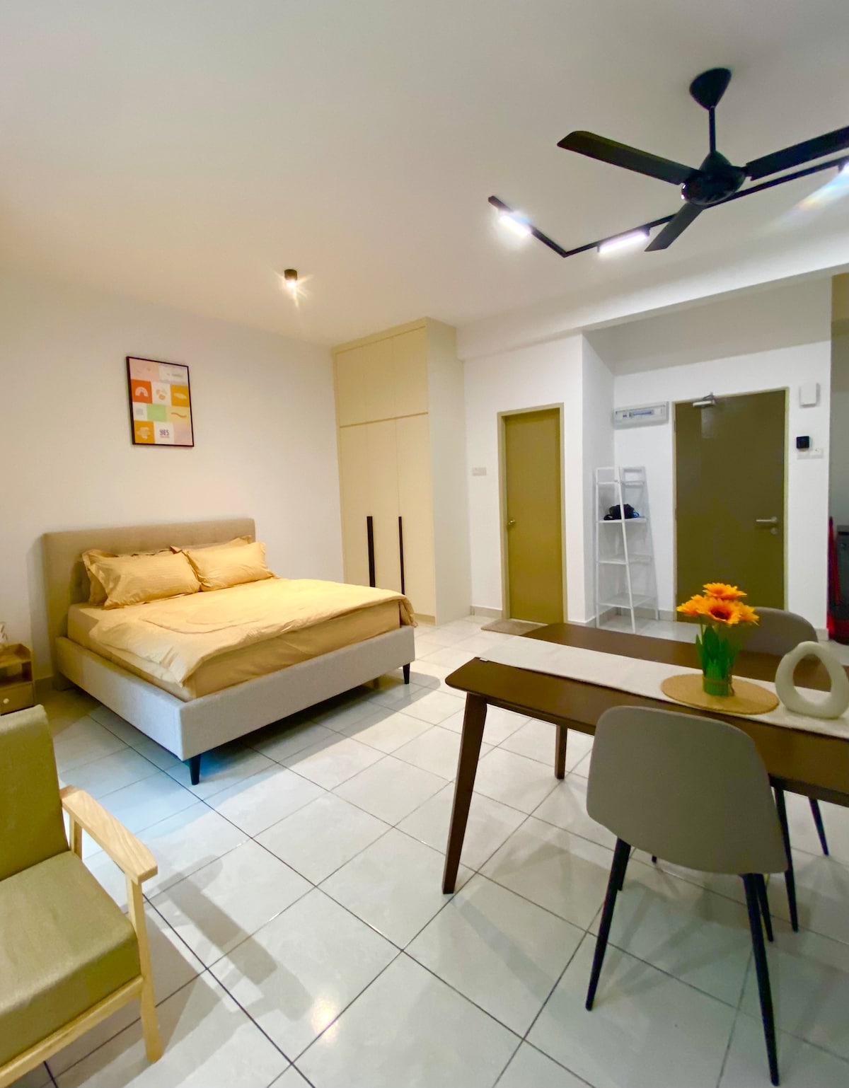 A neutral-toned sleeping area includes a spacious bed with beige bedding. A small dining table with chairs is positioned beside a light-filled window. A comfortable armchair is placed in the corner, while the room features modern lighting and a ceiling fan.