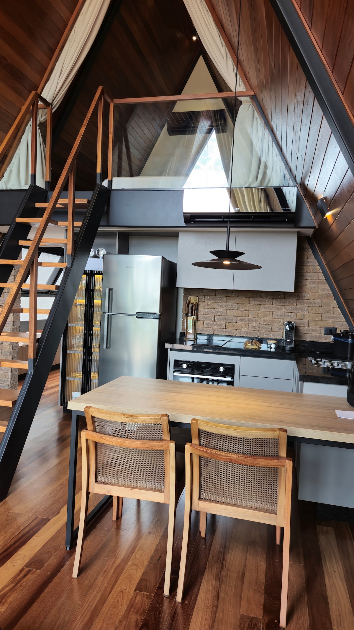 An open kitchen features modern appliances and sleek cabinetry, illuminated by contemporary pendant lighting. A wooden dining table with two chairs is positioned nearby, showcasing the warm tones of the wooden flooring. A staircase leads to the upper level, framed by large windows that enhance natural light.