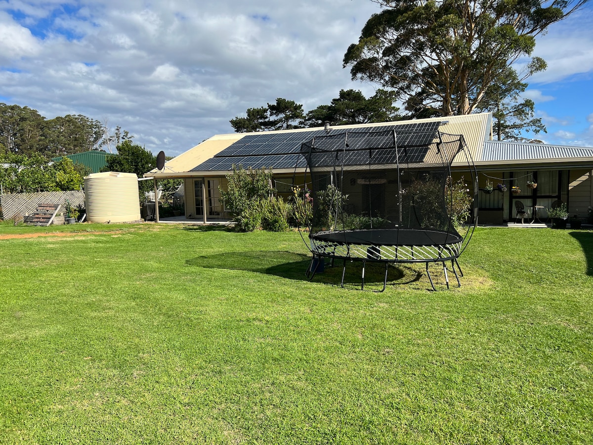 A spacious grassy area features a large trampoline, with solar panels visible on the roof of the nearby house. Natural greenery and garden beds are present, providing a relaxed outdoor space. The area is bathed in soft daylight under a partly cloudy sky.