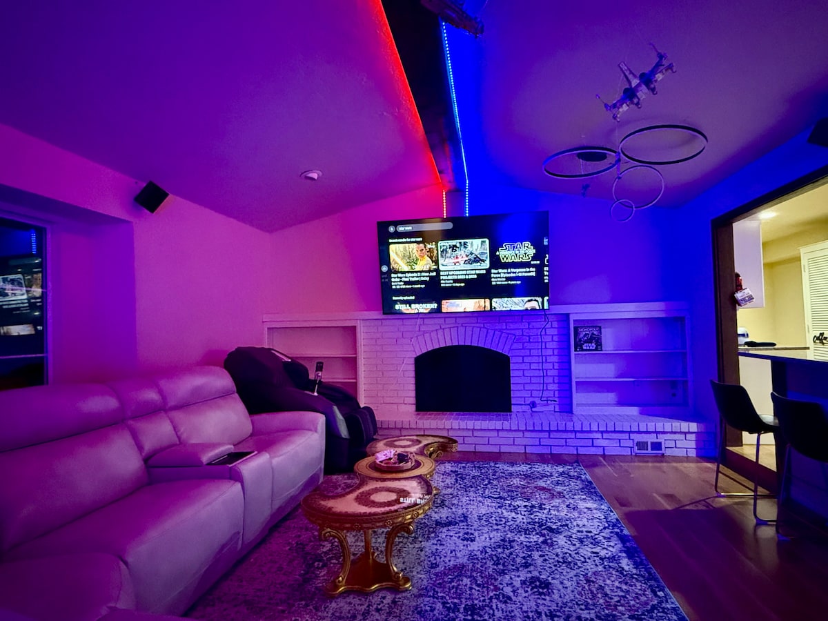 A well-lit living room is presented with colored lights illuminating the ceiling. A large TV is mounted above a brick fireplace, while a sectional sofa is arranged around a decorative coffee table. A soft area rug adds comfort to the hardwood flooring.