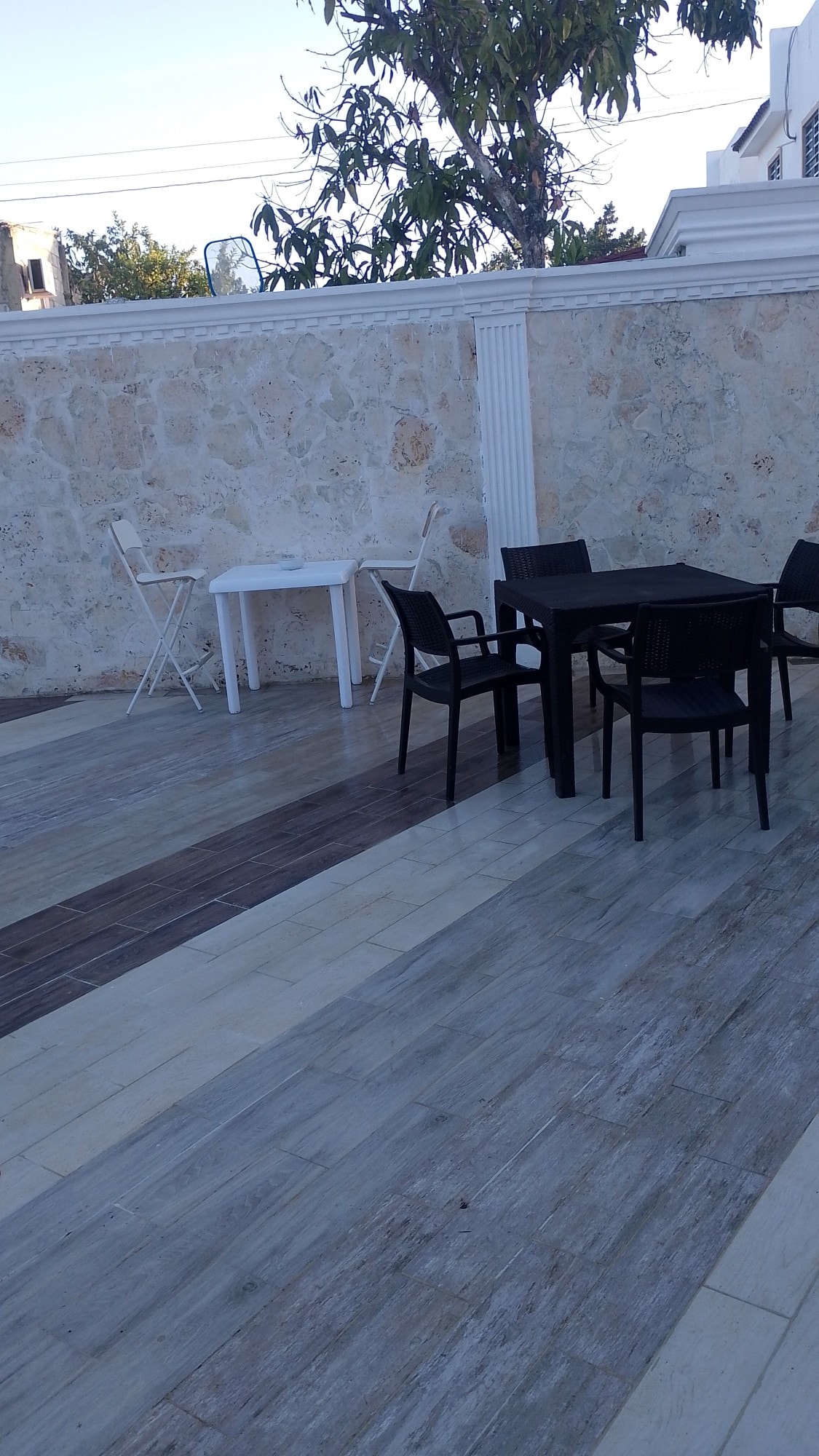 An outdoor seating area features a black rectangular table surrounded by four chairs, set against a natural stone wall. A smaller white table and chair are positioned nearby. The flooring alternates between light and dark tiles, creating a visually interesting surface.