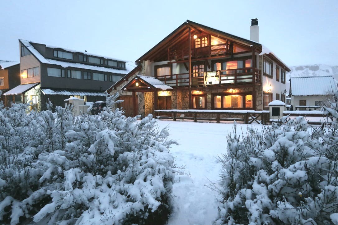 A large wooden house with a stone facade is surrounded by snowy terrain. Warm light emanates from the windows, creating a welcoming ambiance. The structure features multiple levels and balconies, with pine trees and shrubs lightly covered in snow in the foreground.