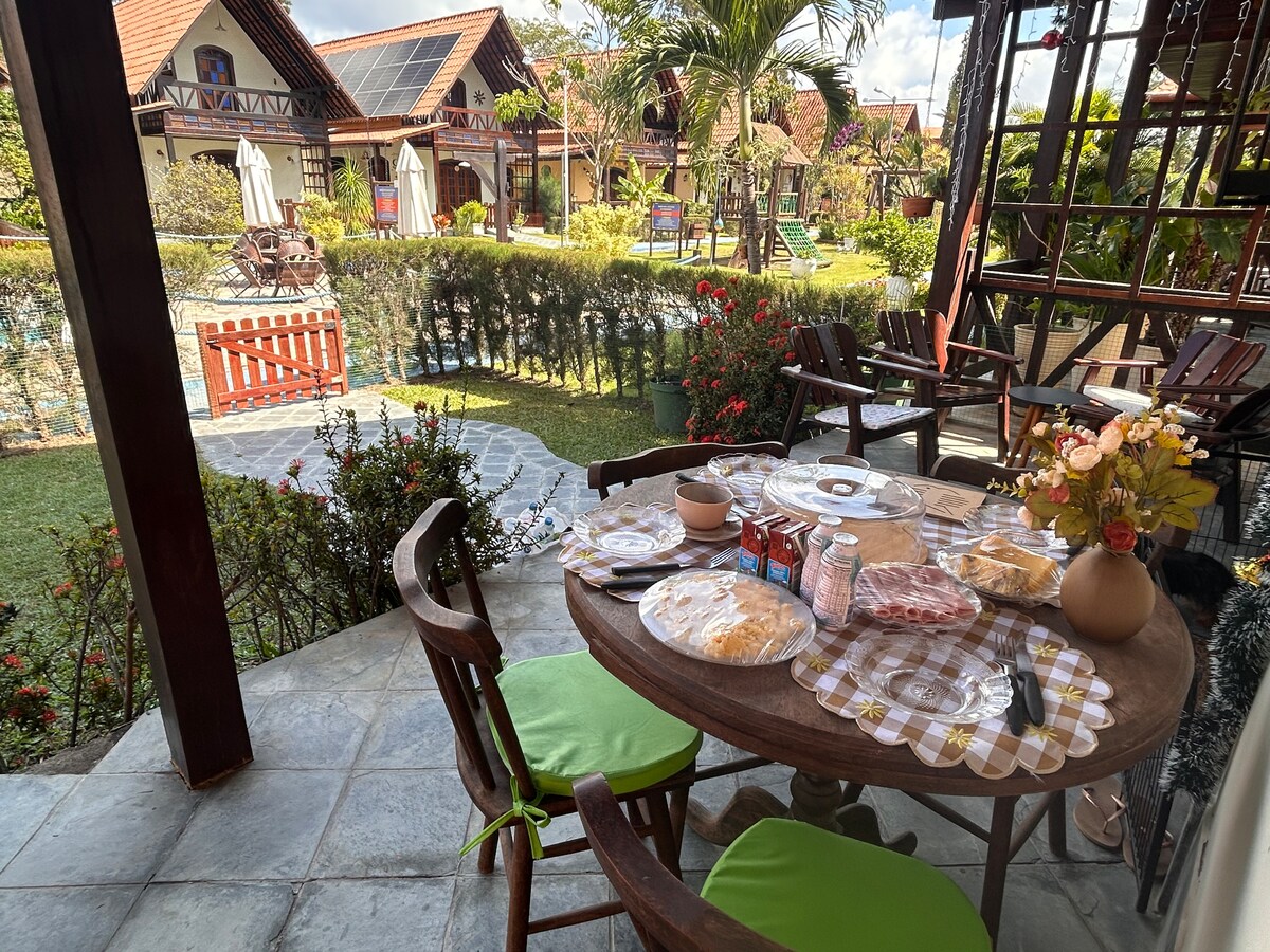 A rustic wooden dining table is set with an assortment of food items, surrounded by chairs with bright green cushions. The outdoor space features a view of well-maintained gardens and nearby chalets, offering a peaceful atmosphere. Natural light illuminates the setting.