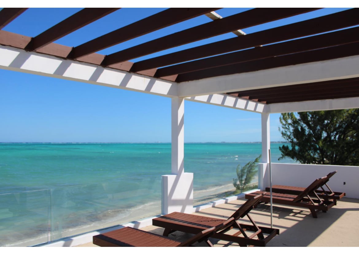A spacious rooftop deck features wooden loungers positioned for ocean views. The open space is framed by a pergola, allowing natural light to fill the area. The turquoise water and sandy beach are visible through the railing, enhancing the outdoor experience.