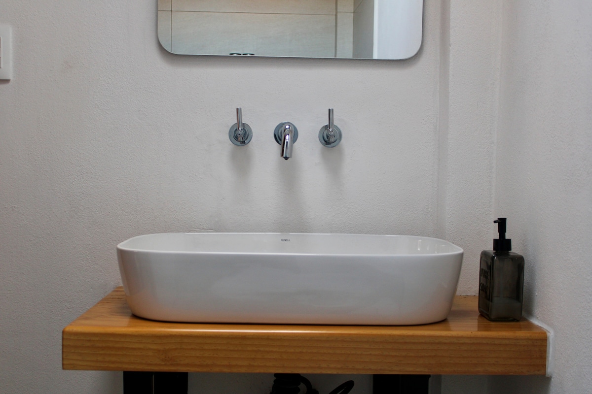 A minimalist bathroom sink area features a simple, modern white basin atop a natural wood countertop. Above the sink, a mirror is mounted on the wall. A sleek soap dispenser is positioned nearby, complementing the clean lines of the space.