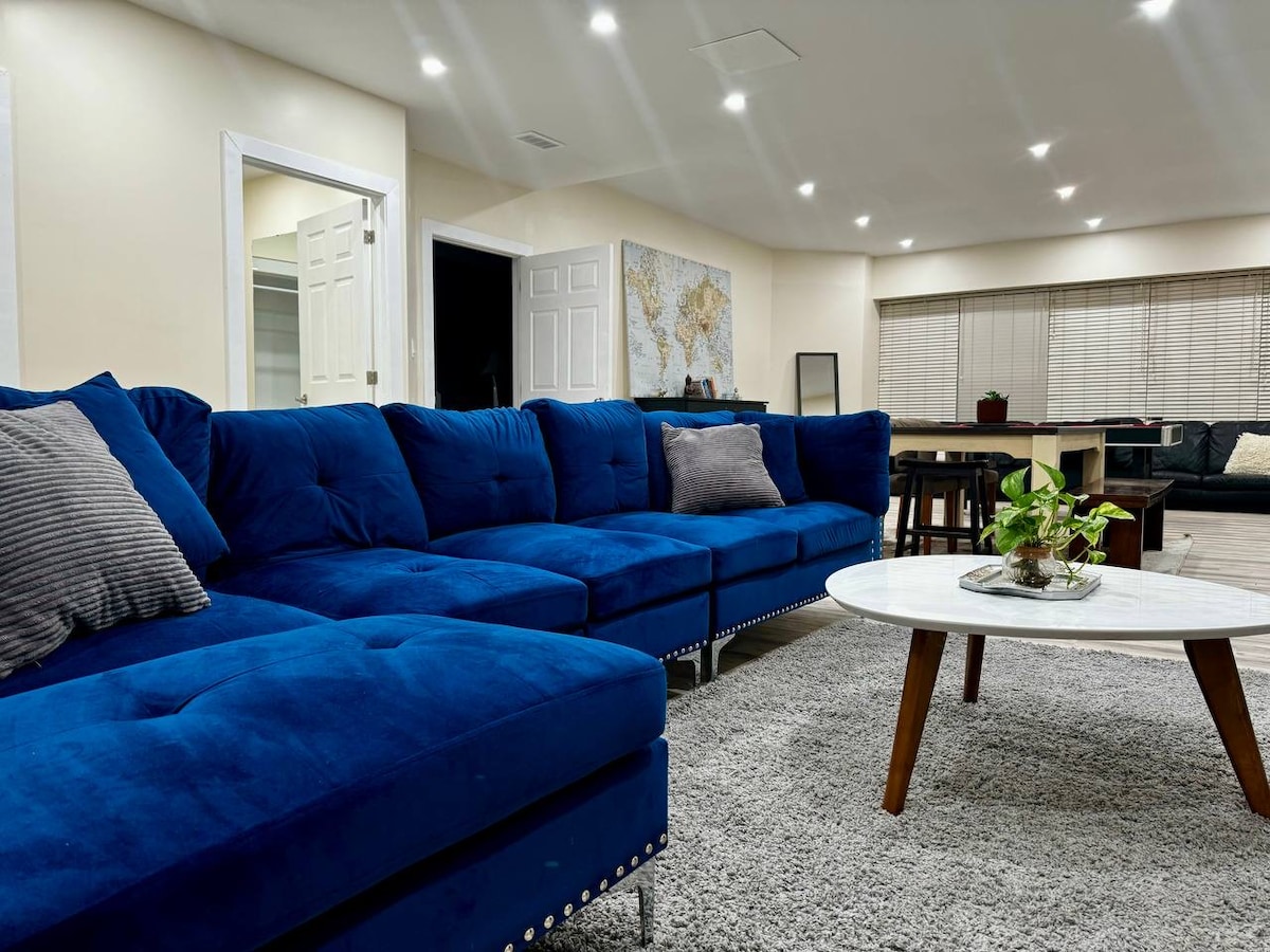 A spacious living area features a large blue sectional sofa adorned with soft cushions. A round coffee table with a plant centerpiece sits on a plush gray rug. Soft lighting illuminates the room, enhancing the welcoming ambiance.