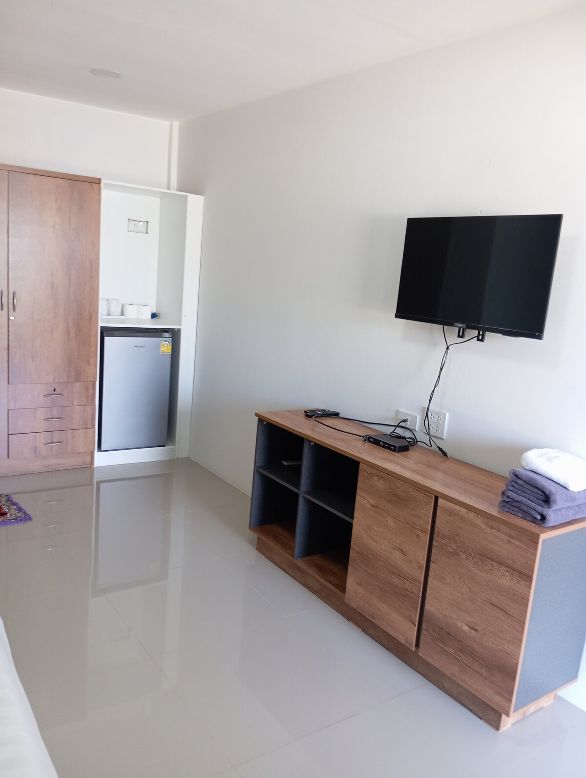 A modern room features light-colored walls and glossy tiled flooring. A wooden cabinet and a television are situated on a low media unit. A small fridge is visible within a built-in kitchenette, complemented by a set of cups arranged on the countertop.