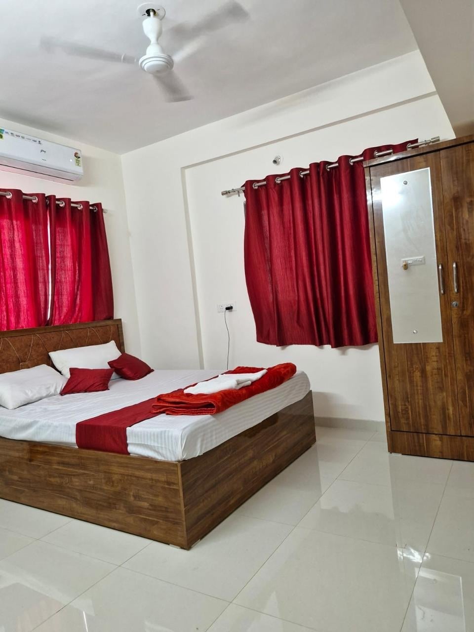 The bedroom features a wooden bed with light bedding arranged neatly. Red curtains adorn the windows, complementing the decorative red accents on the bed. A bedside table and a wardrobe are present, along with a ceiling fan and a wall-mounted air conditioning unit.