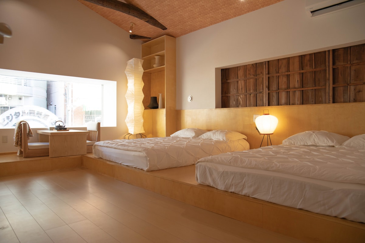 An airy bedroom is presented, featuring two spacious beds positioned on a raised platform. Natural light enters through a large window. A minimalistic desk and chair are located near the window, with a soft lamp offering warm illumination.