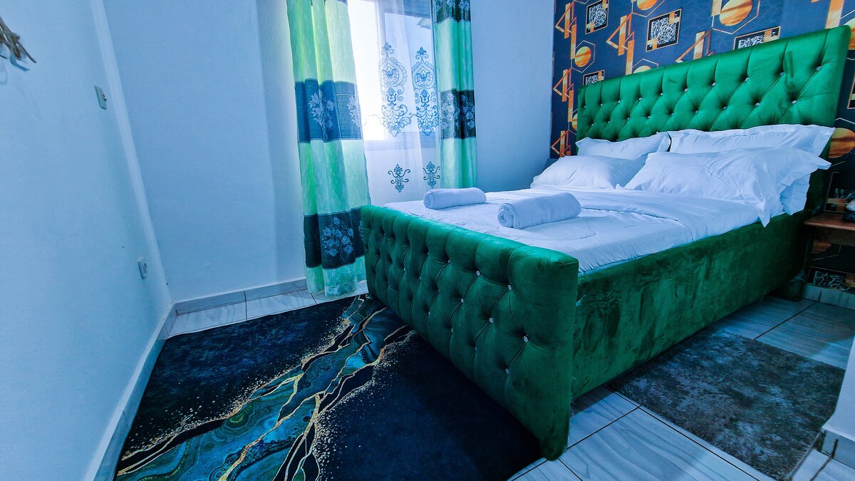 A sleeping area features a plush green upholstered bed with neatly arranged linens and towels. Light is filtered through sheer curtains, illuminating the space. A patterned accent wall and a decorative rug contribute to the room's overall aesthetic.