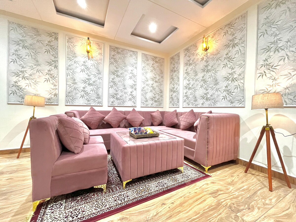 A spacious living area features a modern L-shaped sofa in soft pink upholstery, arranged around an elegant coffee table. Decorative wall panels with a bamboo design contribute to a serene ambiance, complemented by soft lighting from floor lamps placed strategically in the corners.