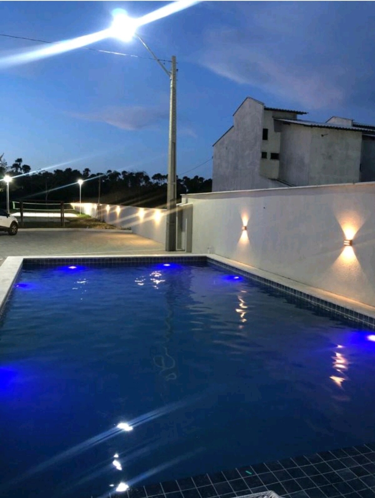 A tranquil pool area is illuminated by soft blue lights, creating a serene atmosphere at dusk. The surrounding walls have built-in lighting, and a white structure is visible in the background, offering a private retreat. The setting reflects a calm and relaxing environment.