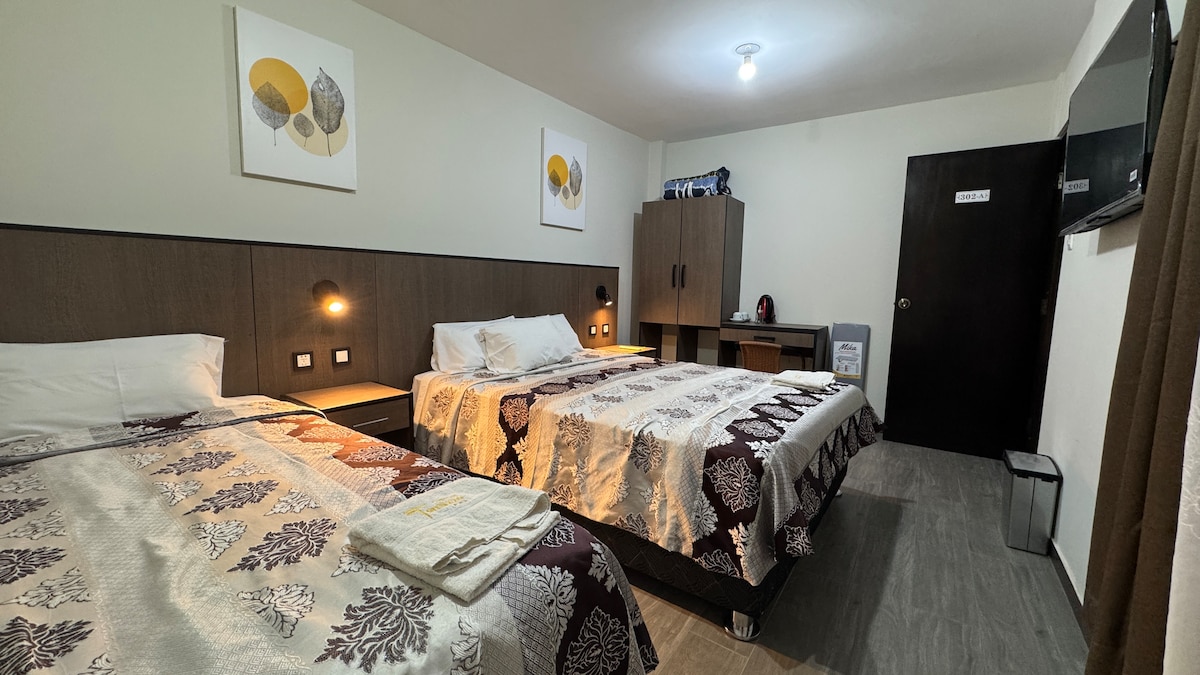 A well-appointed bedroom features two spacious beds with coordinating bedding patterns. Soft lighting from wall fixtures creates a calming environment. A wooden wardrobe and a desk are positioned against the wall, providing functional space. A flat-screen television is mounted for guests' viewing convenience.