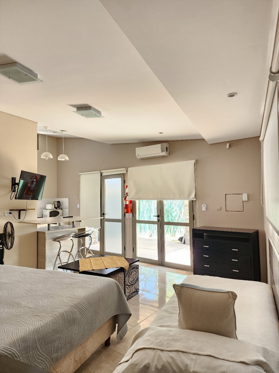 A well-lit sleeping area features a spacious bed covered with a light blanket, accompanied by a neutral-colored cushion. An adjacent seating area with a small table and chairs provides a cozy spot. Large glass doors lead to an outdoor space, allowing natural light to fill the room.