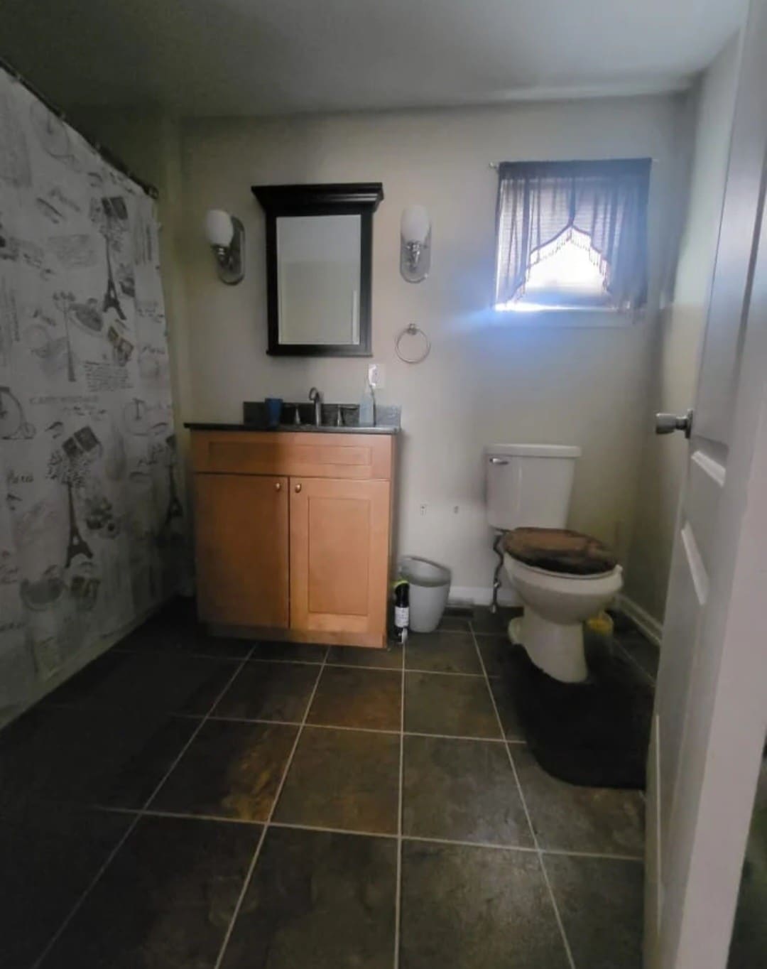 A bathroom includes a modern vanity with a mirror above it, providing ample space for toiletries. A shower area is separated by a decorative curtain. Light is filtered through a small window, illuminating the tiled floor and the toilet positioned nearby.