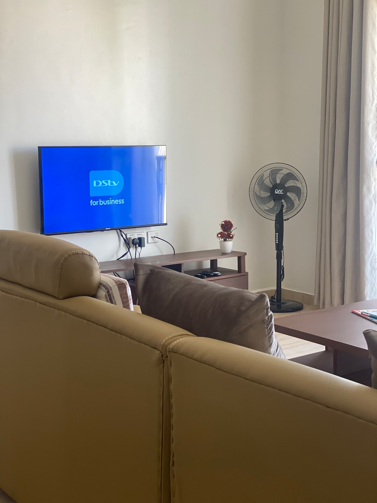A living area features a comfortable seating arrangement with a light-colored sofa. A flat-screen TV is mounted on the wall, displaying the DStv logo. A small table holds decorative items, while a standing fan provides air circulation. Natural light filters through the curtains.