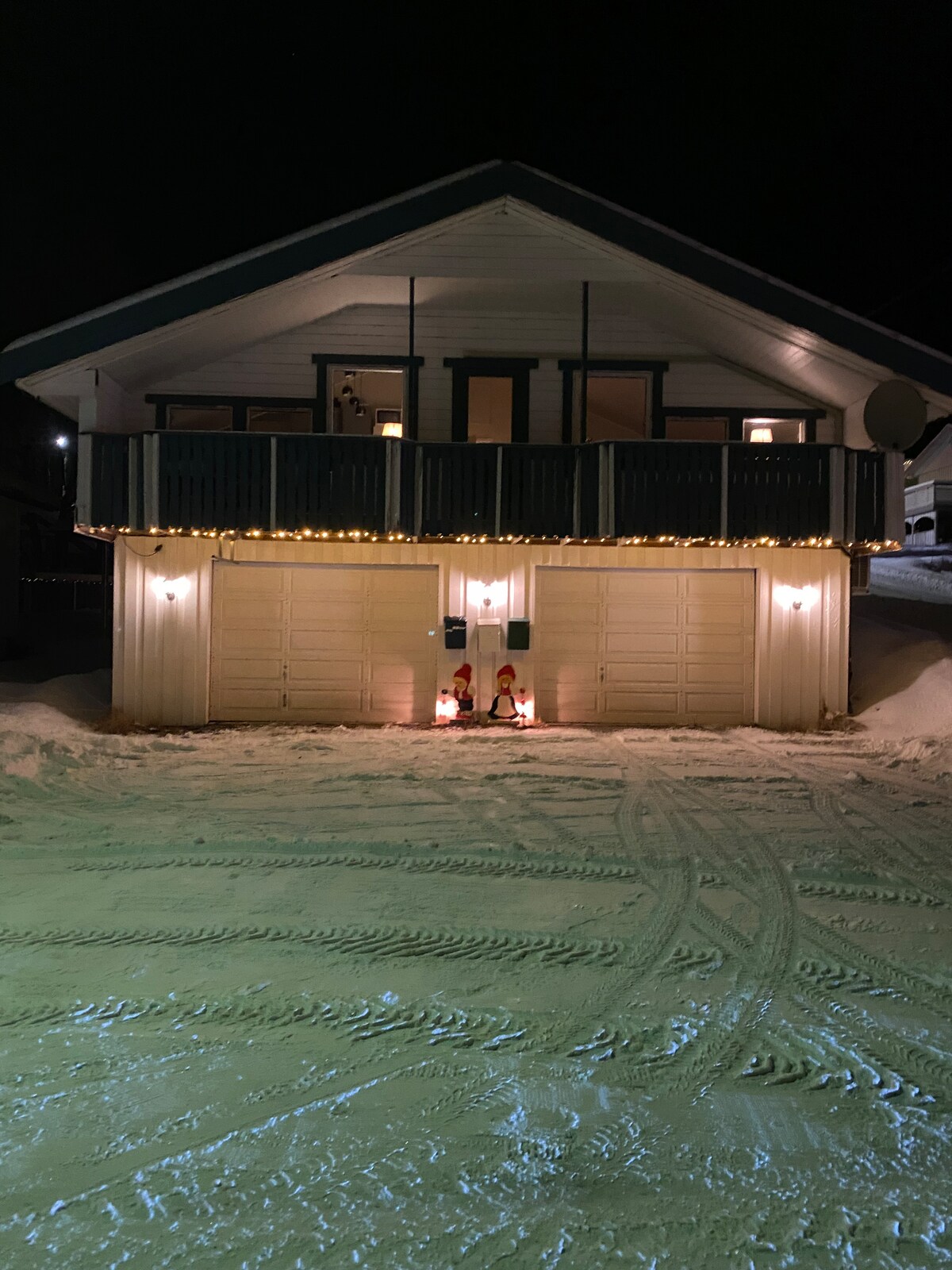 A two-story chalet is presented against a nighttime backdrop, featuring a lit balcony and warm exterior lights. The entrance showcases two decorative figures beside the garages, enhancing the welcoming appearance of the property. Snow coverage adds a serene touch to the surroundings.
