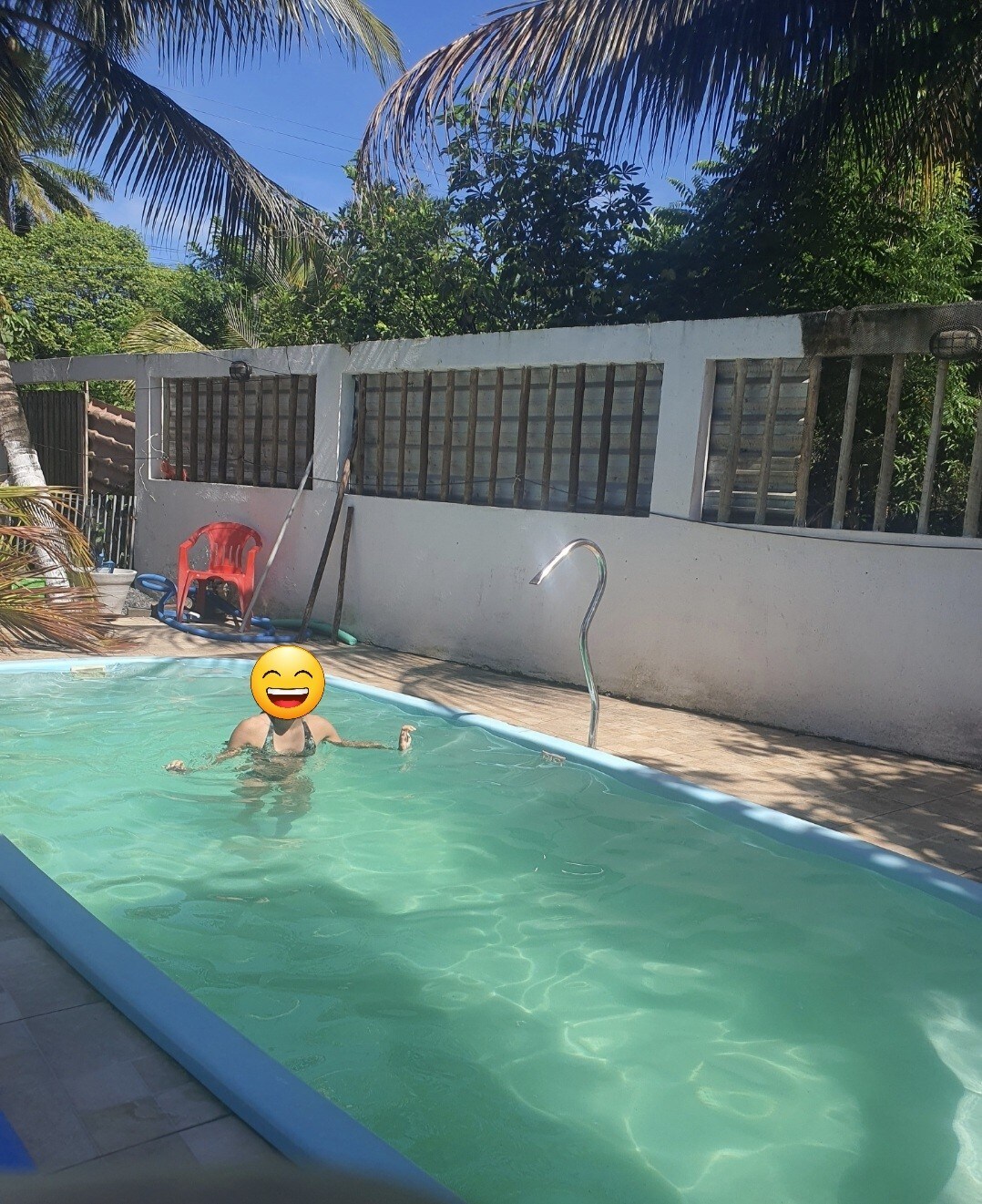 A clear swimming pool is surrounded by a sunlit patio, with a palm tree partially visible. A person is enjoying the water. A red chair is positioned near the pool, and the backdrop features a low wall with several windows.