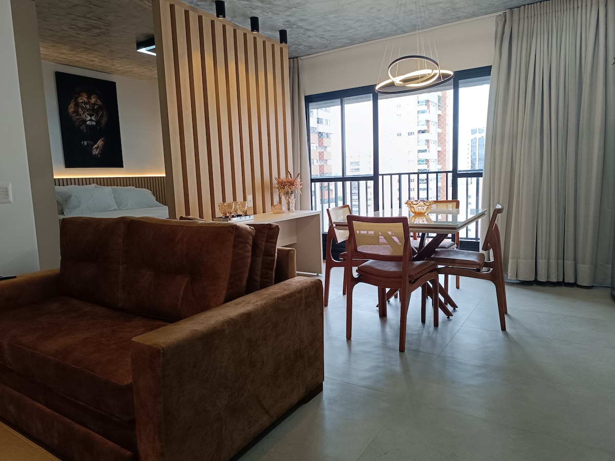 An integrated living space features a comfortable sofa and a dining area with a round table and four chairs. Large windows allow natural light to fill the area, while light-colored curtains add a soft touch. A decorative wall with vertical slats adds dimension to the room.