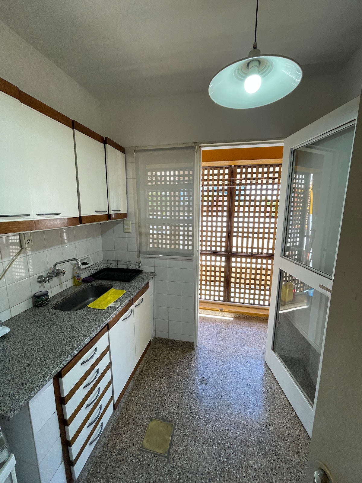 A compact kitchen is shown, featuring light wooden cabinetry and a granite countertop. A sink and stove are installed, with a window providing natural light. A door leads to a balcony, offering additional outdoor space through a slatted screen.