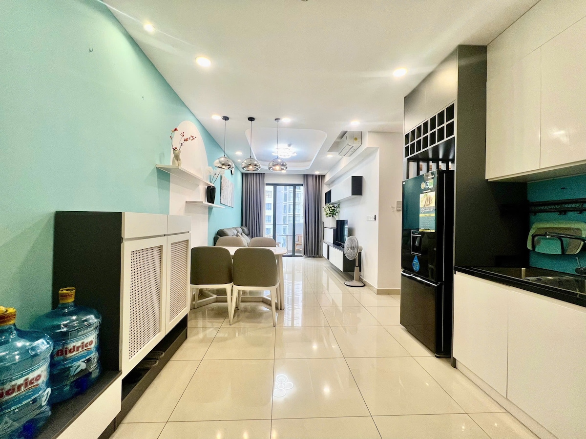 An open living space includes a modern kitchen with black cabinetry and stainless steel appliances. Natural light enters through large windows, illuminating the light-toned tiles. A comfortable seating area features two light grey chairs and a small coffee table, while decorative elements add subtle interest to the room.
