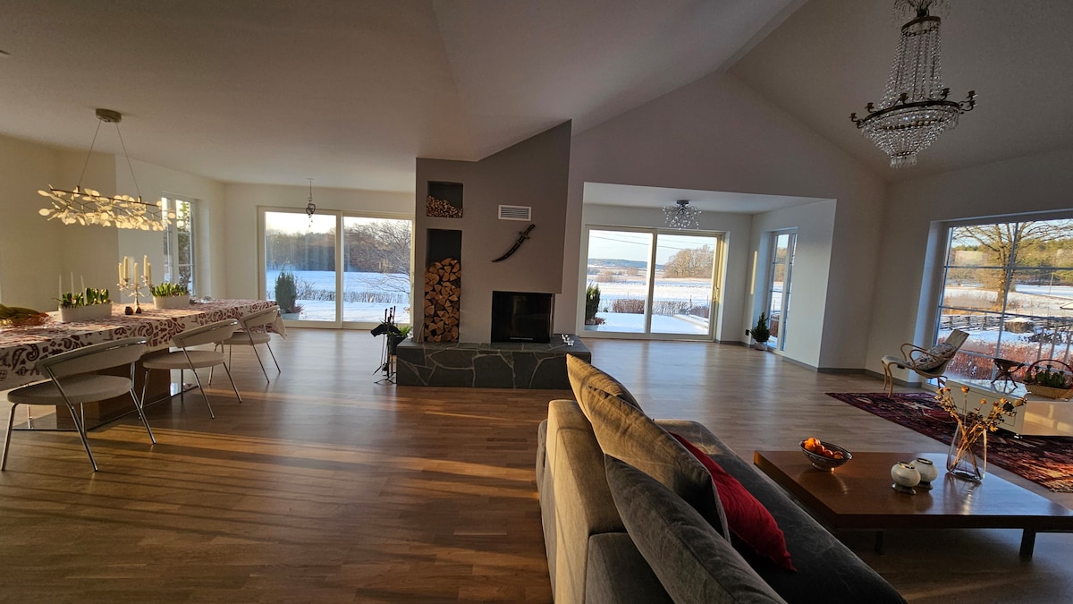 A spacious living area features large windows showcasing views of the serene landscape. A modern fireplace draws attention at the center, surrounded by comfortable seating. A long dining table is set nearby, with subtle lighting above, adding a touch of elegance to the open space.