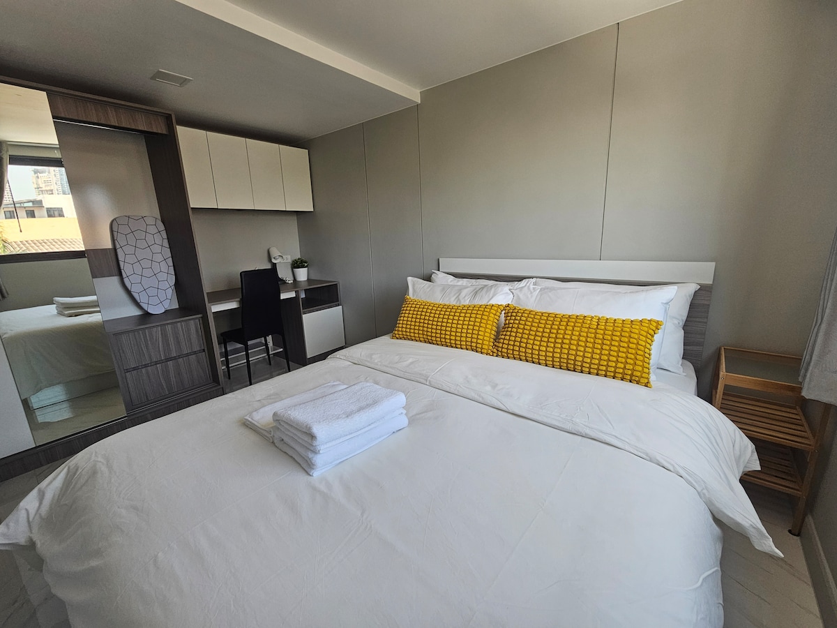 A bright and airy sleeping area features a cozy queen-size bed dressed in crisp white linens. A yellow decorative pillow adds a pop of color. A desk and chair are positioned nearby, alongside a tall mirror and storage cabinets. Natural light enters through a window.