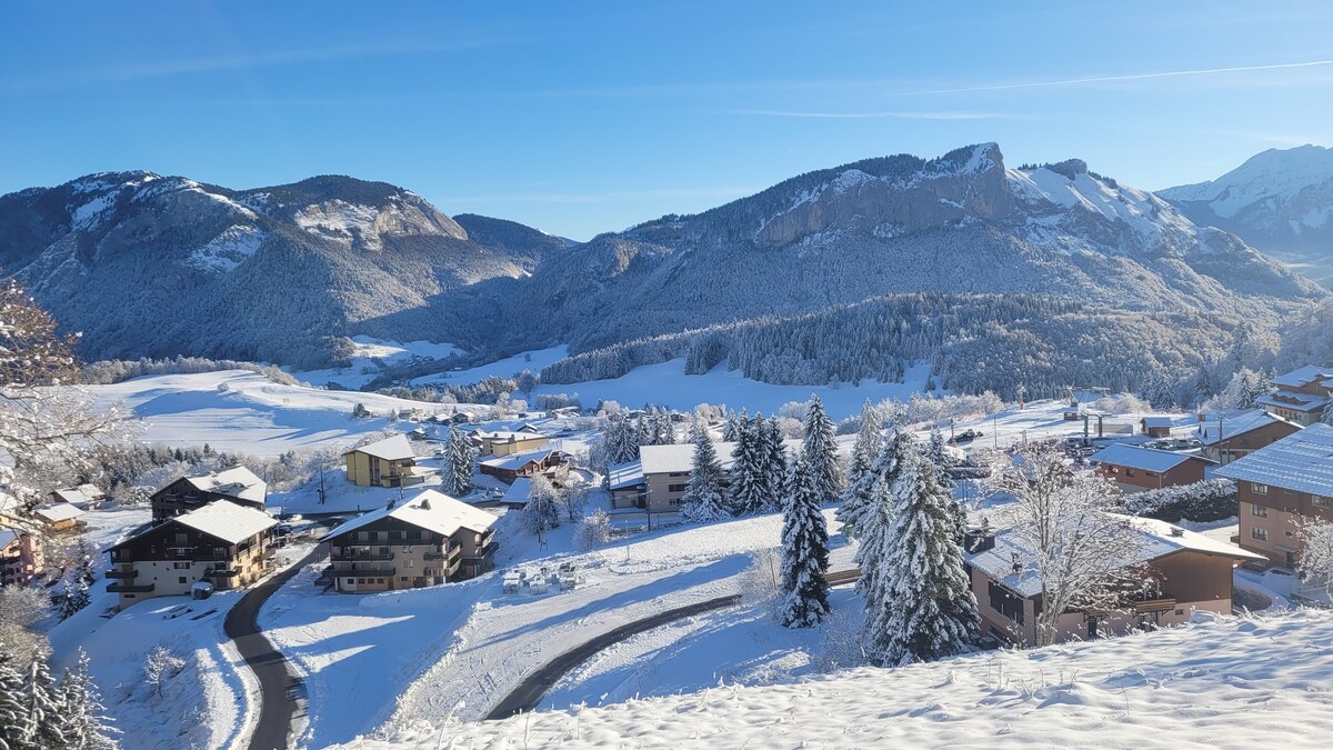 A winter landscape features snow-covered mountains and a serene valley. Residential buildings are visible below, surrounded by snow-laden trees. The clear blue sky enhances the picturesque ambiance, contributing to a tranquil atmosphere.