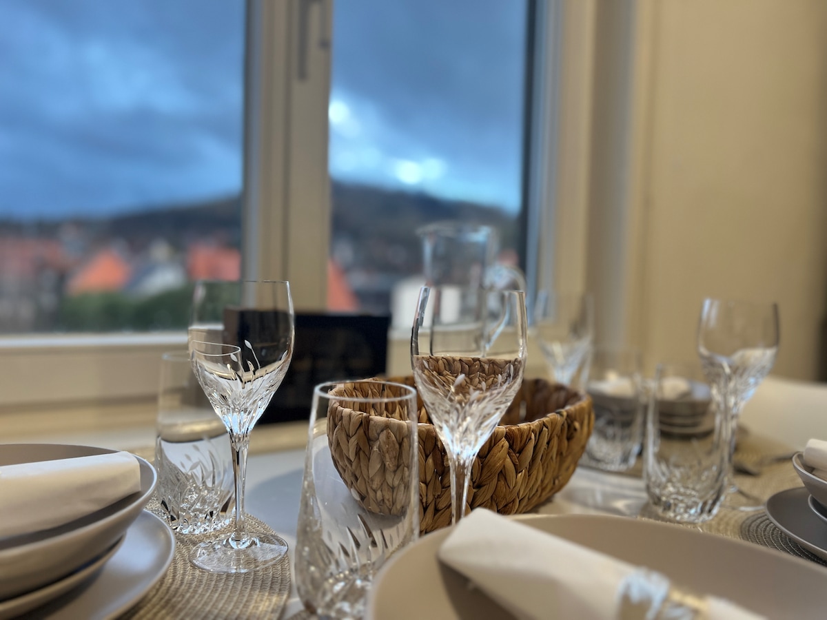 A dining table is set with elegant glassware and plates. Crystal glasses and a woven basket are arranged alongside white napkins, creating a refined dining setting. A view of distant hills and rooftops is visible through a large window, adding a scenic backdrop.